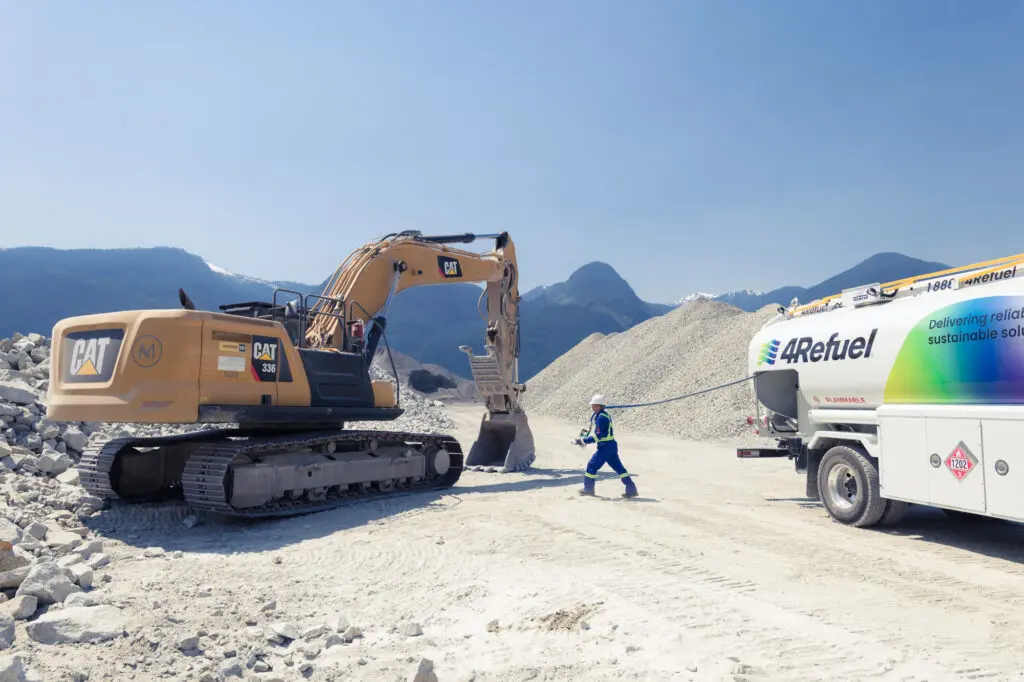 A 4Refuel fuel delivery truck services a CAT 336 excavator at an open-pit quarry or mining site, with a worker in full PPE (white hard hat, blue coveralls, high-visibility vest) walking between the two vehicles. Snow-capped mountains and gravel stockpiles form the backdrop under a clear blue sky.